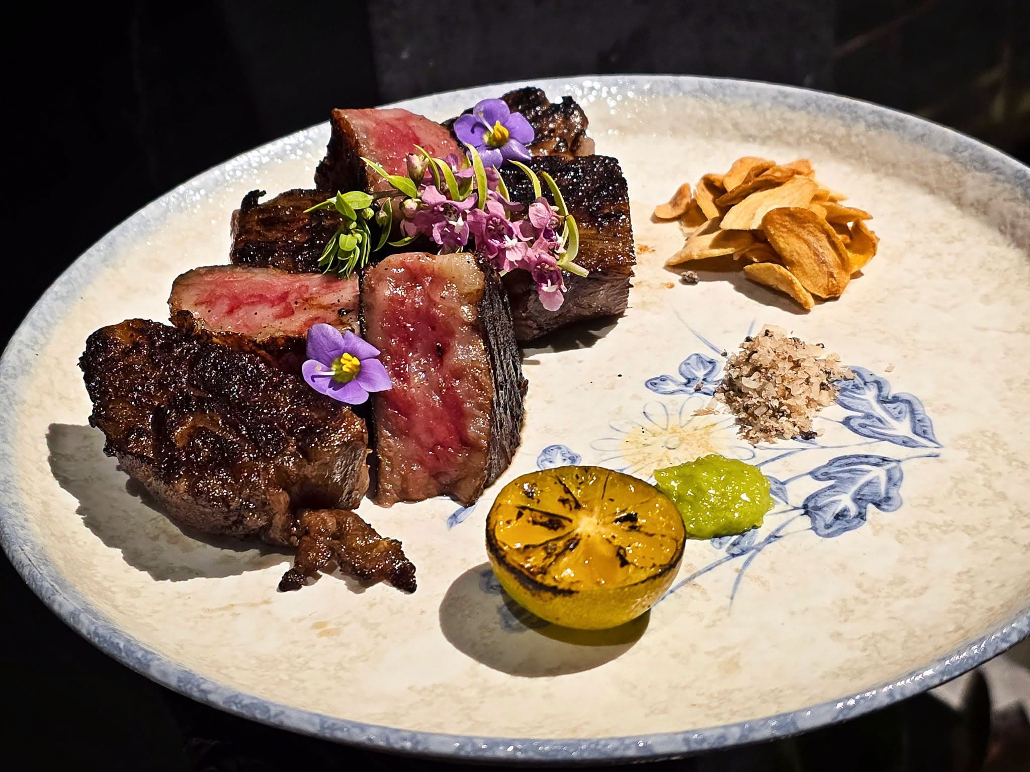 Sliced wagyu beef grilled over fire, plated with garlic crisps, charred citrus and edible flowers