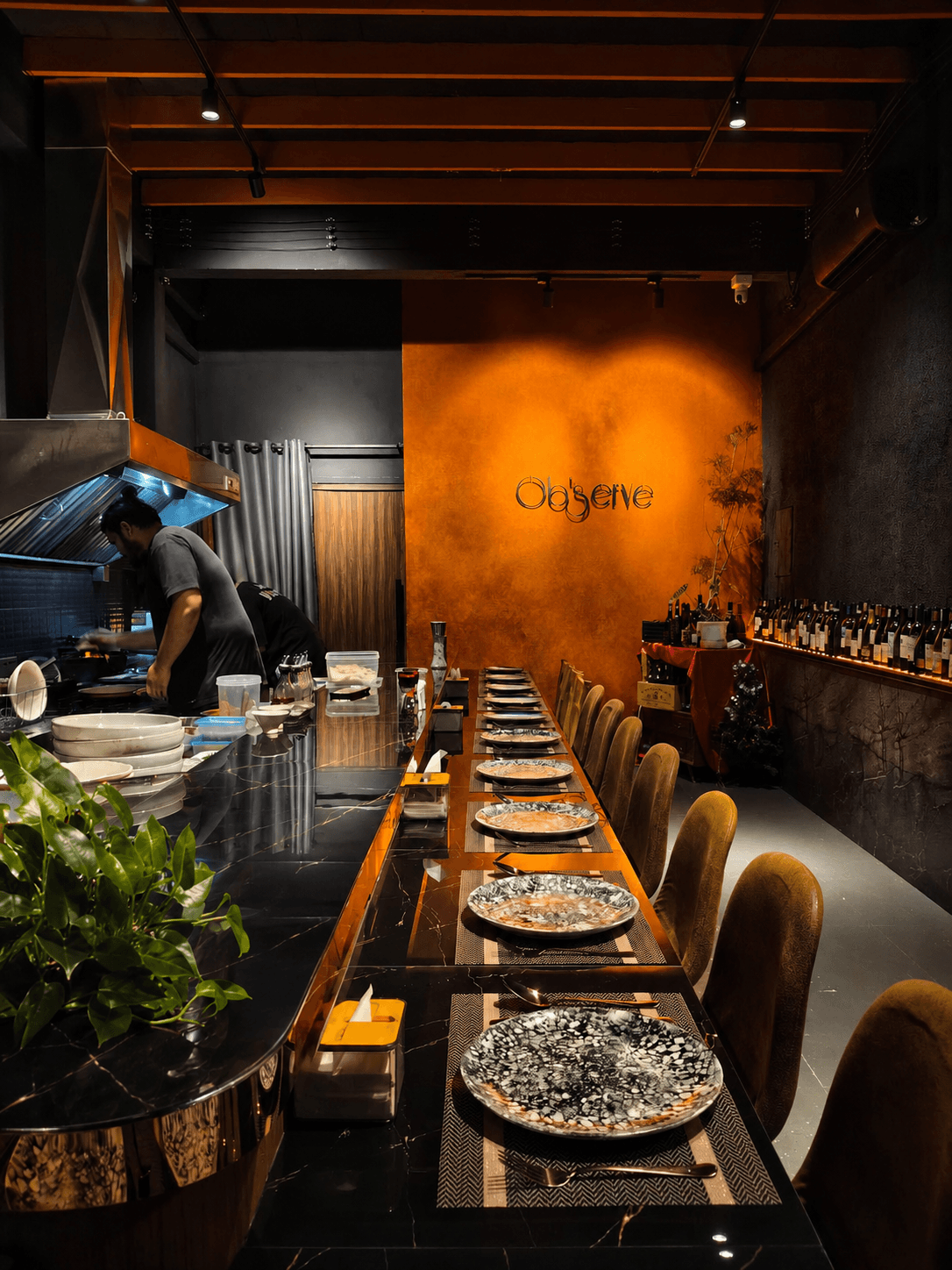 Inside Ob'serve, the long counter set with plates, warm lighting and the Ob'serve sign on the wall as the chefs cook